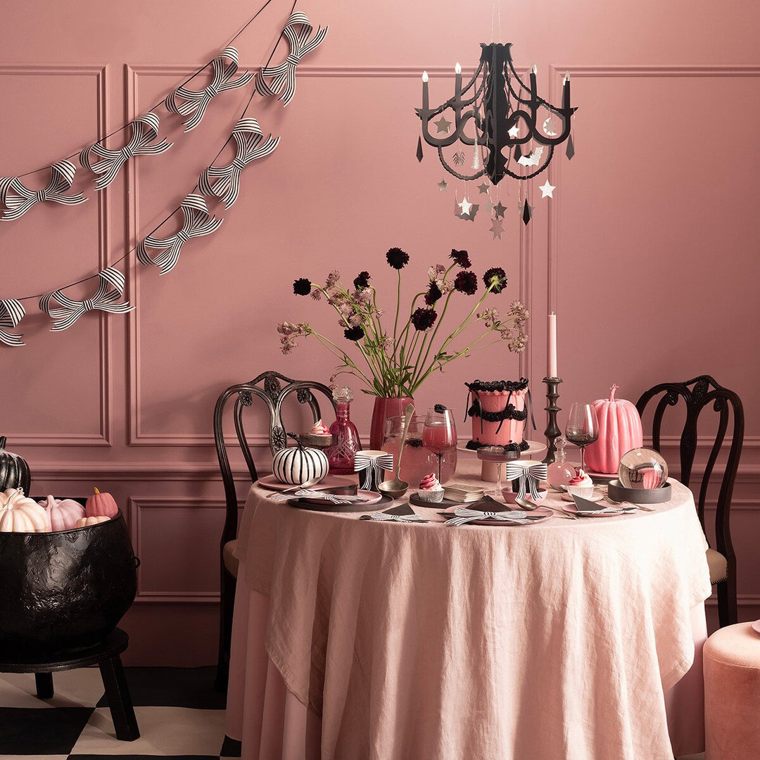 Chic Halloween tableware collection shows a table dressed with a pale pink tablecloth and set with black and white witch hats and napkins with a black and white striped bow garland hanging on the wall.