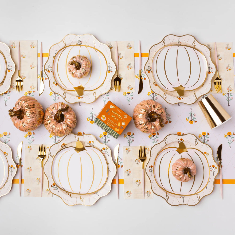 A styled autumn table setting featuring scalloped white plates with gold pumpkin outlines, matching napkins with a floral pattern, gold cutlery, and decorative metallic pumpkins on a light gray background.