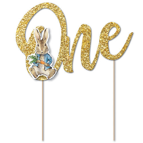 A glittery gold 'One' birthday cake topper with a Peter Rabbit illustration on the letter 'O', mounted on two wooden sticks against a plain white background.