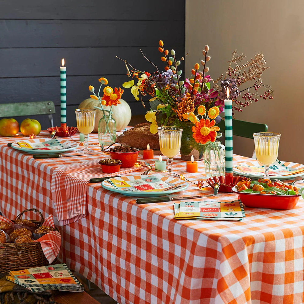 Autumn table styling with orange and white gingham cloth and brightly coloured tableware, floral arrangements in autumnal colours and lit candles. 